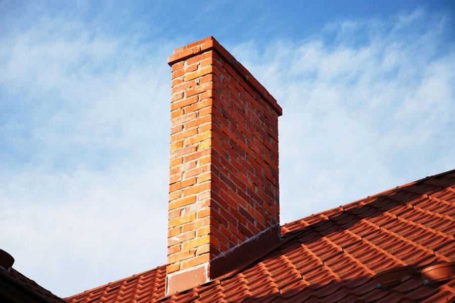 A tall, rectangular brick chimney rises from a sloped red-tiled roof, showcasing well-maintained brickwork against a blue sky with wispy white clouds.