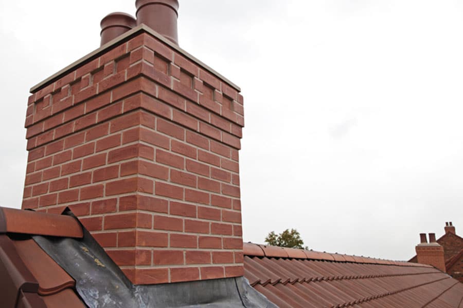 A close-up view of a red brick chimney with two flues on a sloped, tiled roof against a cloudy sky highlights the quality brickwork. A small section of another roof and chimney is visible in the background.