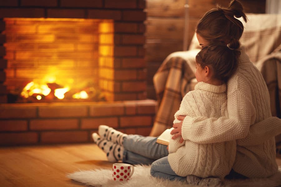 A woman and child in cozy sweaters sit by a lit brick fireplace, hugging as they enjoy the essential warmth of winter. A polka dot mug rests on a fluffy rug nearby, adding comfort and charm—reminding us of the value of chimney inspections.