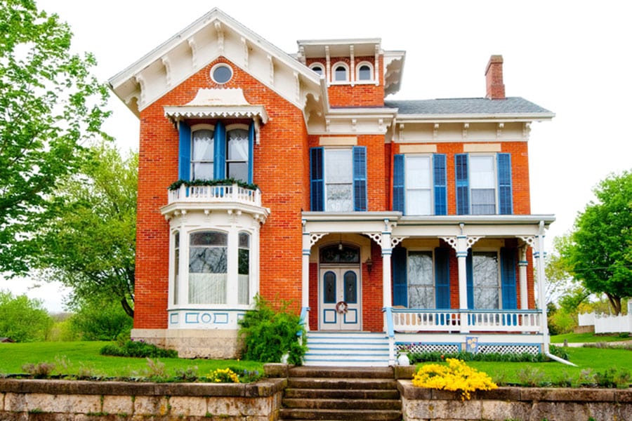 A red brick Victorian-style house with blue trim, white decorative woodwork, detailed brickwork, tall windows, a wraparound porch, and a bay window, surrounded by green trees and shrubs on a landscaped lawn.