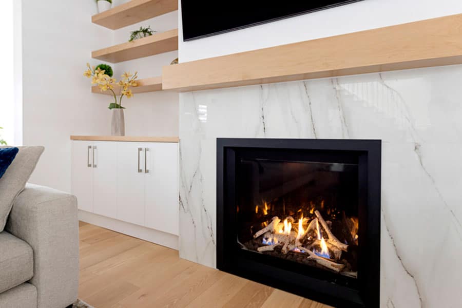 A modern living room with a marble fireplace, burning firewood—essential for cozy winter nights—light wood shelves with decor, white cabinets with silver handles, and a beige sofa partially visible on the left.