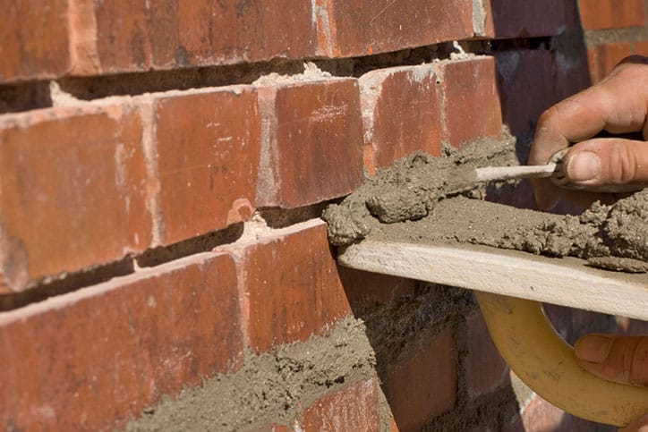 A close-up of a person’s hand applying mortar between red bricks with a trowel during brick wall construction or Brick Restoration in Metro Detroit.